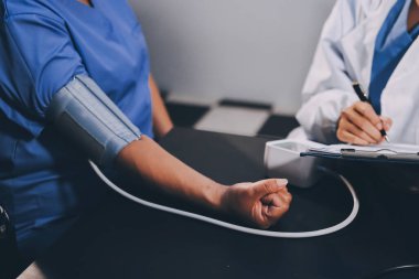 Hastanın tansiyonunu ölçen bir doktor. Hastanın kolunda tansiyon aleti var. Doktor da tansiyon monitörü kullanıyor..