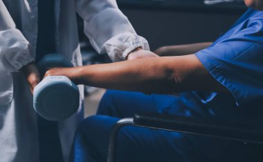 Evde dambıl kullanarak fizyoterapist eğitimi alan yaşlı bir kadın. Terapist, huzurevi egzersizlerinde yaşlı kadına yardım ediyor. Yaşlı bir hasta kollarını uzatmış halterler kullanıyor..