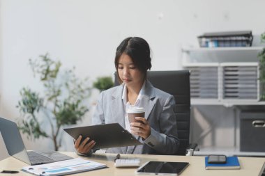 Başarılı Asyalı iş kadını dizüstü bilgisayarla gülümseyip ofisinde kahve fincanı tutuyor. Güvenilir Asyalı iş kadını ofiste mutlu mesut oturuyor..