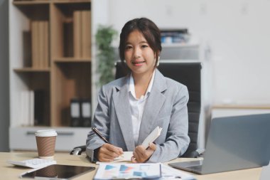 Başarılı Asyalı iş kadını dizüstü bilgisayarla gülümseyip ofisinde kahve fincanı tutuyor. Güvenilir Asyalı iş kadını ofiste mutlu mesut oturuyor..