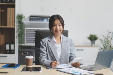 Başarılı Asyalı iş kadını dizüstü bilgisayarla gülümseyip ofisinde kahve fincanı tutuyor. Güvenilir Asyalı iş kadını ofiste mutlu mesut oturuyor..