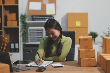 Girişim KOBİ küçük işletme girişimcisi önlük giyen Asyalı kadın dizüstü bilgisayar ve kutu kullanarak çevrimiçi müşterilere, çevrimiçi sme iş fikirlerine satış hazırlamak için siparişleri alır ve gözden geçirir.