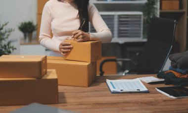 Girişim KOBİ küçük işletme girişimcisi önlük giyen Asyalı kadın dizüstü bilgisayar ve kutu kullanarak çevrimiçi müşterilere, çevrimiçi sme iş fikirlerine satış hazırlamak için siparişleri alır ve gözden geçirir.