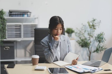 Başarılı Asyalı iş kadını dizüstü bilgisayarla gülümseyip ofisinde kahve fincanı tutuyor. Güvenilir Asyalı iş kadını ofiste mutlu mesut oturuyor..