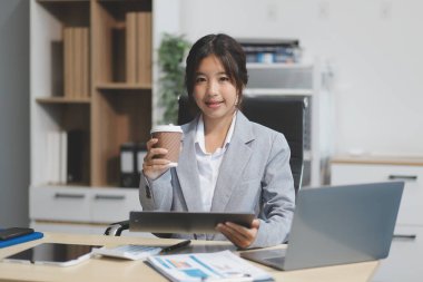 Başarılı Asyalı iş kadını dizüstü bilgisayarla gülümseyip ofisinde kahve fincanı tutuyor. Güvenilir Asyalı iş kadını ofiste mutlu mesut oturuyor..