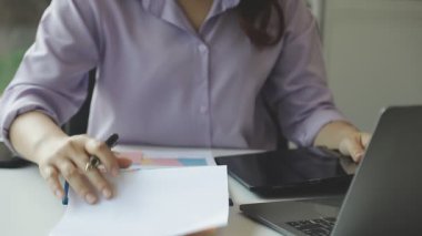 Genç iş kadını modern ofiste otururken dizüstü bilgisayar kullanıyor ve elinde kalem tutuyor..