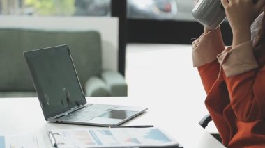 Genç iş kadını elinde kahve fincanıyla ofiste oturuyor ve bilgisayar kullanıyor.