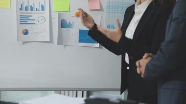 Takım elbiseli genç Asyalı işadamı ve siyah takım elbiseli adam ofisteki meslektaşlarıyla birlikte grafiği işaret ediyor.