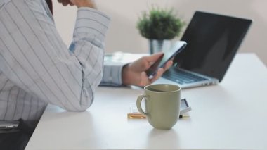 İş adamı dizüstü bilgisayar kullanıyor, akıllı telefonunu elinde tutuyor ve ofiste kahve içiyor..