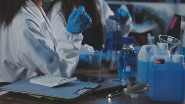 Mavi eldivenli ve gözlüklü laboratuvar asistanı. Masada sıvı var. Kapat.