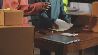 Adam dizüstü bilgisayarda çalışıyor ve depoda karton kutu paketliyor.