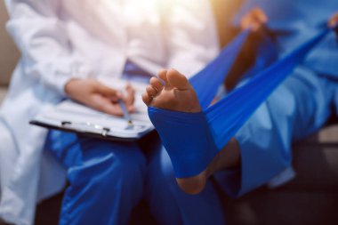 Kendine güvenen fizyoterapist, hastanın bacağını klinik odasında gerdirerek direnç bandı kullanmasına yardım ediyor..