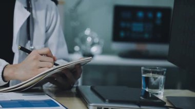 Genç erkek doktor, klinikteki masada otururken dizüstü bilgisayar kullanıyor ve not defterine notlar yazıyor.