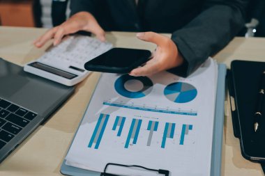 Genç, gülümseyen Asyalı iş kadını cep telefonu kullanırken omzunda bir ceket tutuyor, şık ve modern bir ofis ortamında kendine güvenerek duruyor.