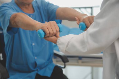 Yaşlı Asyalı adam hemşirenin desteğiyle fizyoterapiste gidiyor. Oturma odasındaki kanepede oturan yaşlı erkek hasta bakıcısı için halterli egzersiz yapıyor..