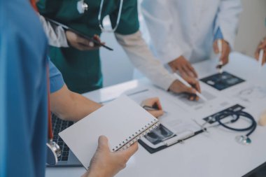 Çok ırklı tıbbi ekip, beyaz laboratuvar önlüklü ve ameliyat önlüklü doktorlarla toplantı yapıyor. Hasta kayıtlarını tartışıyorlar.