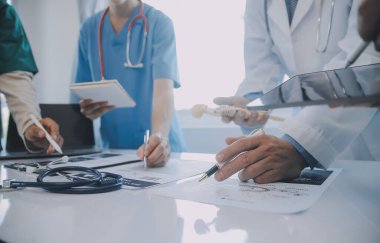 Çok ırklı tıbbi ekip, beyaz laboratuvar önlüklü ve ameliyat önlüklü doktorlarla toplantı yapıyor. Hasta kayıtlarını tartışıyorlar.