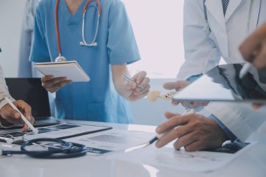 Çok ırklı tıbbi ekip, beyaz laboratuvar önlüklü ve ameliyat önlüklü doktorlarla toplantı yapıyor. Hasta kayıtlarını tartışıyorlar.