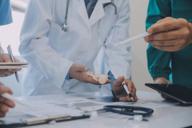 Çok ırklı tıbbi ekip, beyaz laboratuvar önlüklü ve ameliyat önlüklü doktorlarla toplantı yapıyor. Hasta kayıtlarını tartışıyorlar.