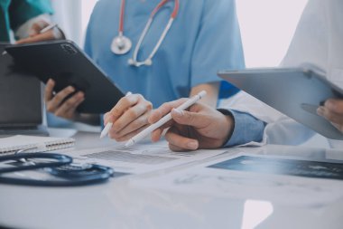 Çok ırklı tıbbi ekip, beyaz laboratuvar önlüklü ve ameliyat önlüklü doktorlarla toplantı yapıyor. Hasta kayıtlarını tartışıyorlar.