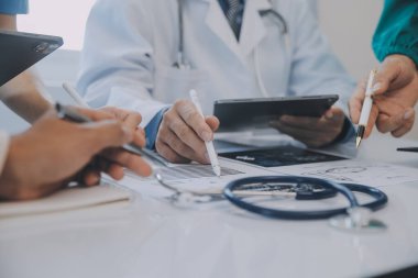 Çok ırklı tıbbi ekip, beyaz laboratuvar önlüklü ve ameliyat önlüklü doktorlarla toplantı yapıyor. Hasta kayıtlarını tartışıyorlar.