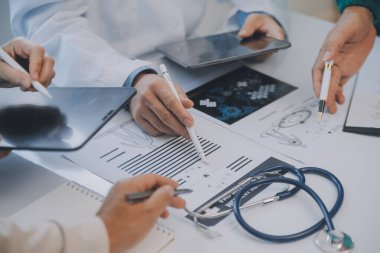 Çok ırklı tıbbi ekip, beyaz laboratuvar önlüklü ve ameliyat önlüklü doktorlarla toplantı yapıyor. Hasta kayıtlarını tartışıyorlar.