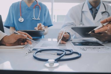 Çok ırklı tıbbi ekip, beyaz laboratuvar önlüklü ve ameliyat önlüklü doktorlarla toplantı yapıyor. Hasta kayıtlarını tartışıyorlar.