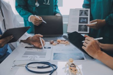 Çok ırklı tıbbi ekip, beyaz laboratuvar önlüklü ve ameliyat önlüklü doktorlarla toplantı yapıyor. Hasta kayıtlarını tartışıyorlar.