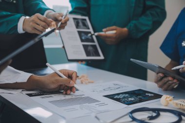 Çok ırklı tıbbi ekip, beyaz laboratuvar önlüklü ve ameliyat önlüklü doktorlarla toplantı yapıyor. Hasta kayıtlarını tartışıyorlar.