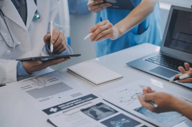 Çok ırklı tıbbi ekip, beyaz laboratuvar önlüklü ve ameliyat önlüklü doktorlarla toplantı yapıyor. Hasta kayıtlarını tartışıyorlar.