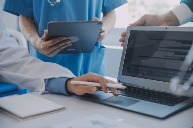 Çok ırklı tıbbi ekip, beyaz laboratuvar önlüklü ve ameliyat önlüklü doktorlarla toplantı yapıyor. Hasta kayıtlarını tartışıyorlar.