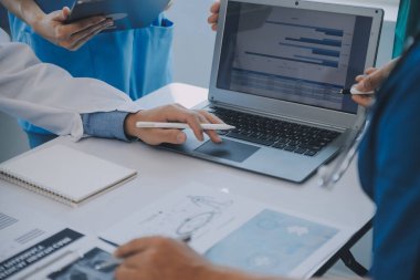 Çok ırklı tıbbi ekip, beyaz laboratuvar önlüklü ve ameliyat önlüklü doktorlarla toplantı yapıyor. Hasta kayıtlarını tartışıyorlar.