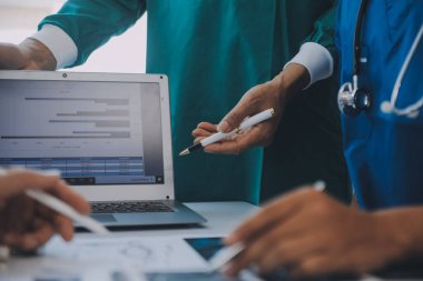 Çok ırklı tıbbi ekip, beyaz laboratuvar önlüklü ve ameliyat önlüklü doktorlarla toplantı yapıyor. Hasta kayıtlarını tartışıyorlar.