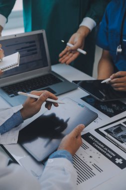 Çok ırklı tıbbi ekip, beyaz laboratuvar önlüklü ve ameliyat önlüklü doktorlarla toplantı yapıyor. Hasta kayıtlarını tartışıyorlar.