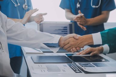 Çok ırklı tıbbi ekip, beyaz laboratuvar önlüklü ve ameliyat önlüklü doktorlarla toplantı yapıyor. Hasta kayıtlarını tartışıyorlar.