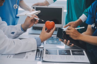 Çok ırklı tıbbi ekip, beyaz laboratuvar önlüklü ve ameliyat önlüklü doktorlarla toplantı yapıyor. Hasta kayıtlarını tartışıyorlar.