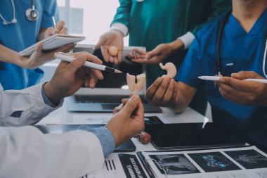 Çok ırklı tıbbi ekip, beyaz laboratuvar önlüklü ve ameliyat önlüklü doktorlarla toplantı yapıyor. Hasta kayıtlarını tartışıyorlar.