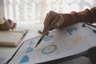 İşadamı elinde kalemle kağıt üzerinde yazı yazıyor. Ahşap masa üzerinde de mali tabloları var. ofis ve iş konsepti.