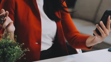 Beyaz takım elbiseli ve kırmızı elbiseli genç bir iş kadını modern bir ofiste tablet ve cep telefonuyla çalışıyor.