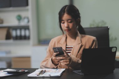 Asyalı Kadın girişimci ofiste işleriyle meşgul. Genç Asyalı kadın masasında bilgisayar başında çalışırken akıllı telefondan ya da cep telefonundan konuşuyor..