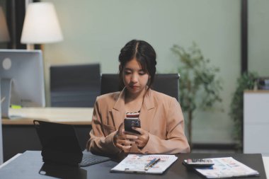 Asyalı Kadın girişimci ofiste işleriyle meşgul. Genç Asyalı kadın masasında bilgisayar başında çalışırken akıllı telefondan ya da cep telefonundan konuşuyor..