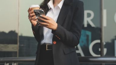 Siyah takım elbiseli iş kadını elinde akıllı telefon ve kahve fincanı tutuyor.