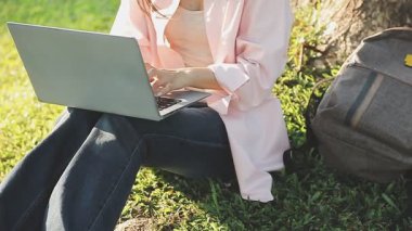 Laptop ve kulaklıklarla yeşil çimen parkında oturan genç bir kadın.