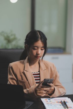 Asyalı Kadın girişimci ofiste işleriyle meşgul. Genç Asyalı kadın masasında bilgisayar başında çalışırken akıllı telefondan ya da cep telefonundan konuşuyor..