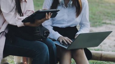 İki genç iş kadını ahşap bir bankta oturuyor ve dizüstü bilgisayar kullanıyorlar..