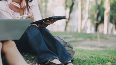 Genç Asyalı kadın doğadaki çimlerin üzerinde dizüstü bilgisayar ve defter kullanıyor..