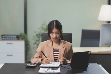 Telework konsepti - Asyalı iş kadını video toplantısına katılmak için dizüstü bilgisayar kullanır ve evdeki dijital tabletine notlar yazar
