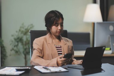 Asyalı Kadın girişimci ofiste işleriyle meşgul. Genç Asyalı kadın masasında bilgisayar başında çalışırken akıllı telefondan ya da cep telefonundan konuşuyor..