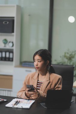Asyalı Kadın girişimci ofiste işleriyle meşgul. Genç Asyalı kadın masasında bilgisayar başında çalışırken akıllı telefondan ya da cep telefonundan konuşuyor..