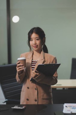 Mutlu Asyalı iş kadını projesi için çalışıyor ve çalışırken kahve içiyor, ofis masasında oturuyor..